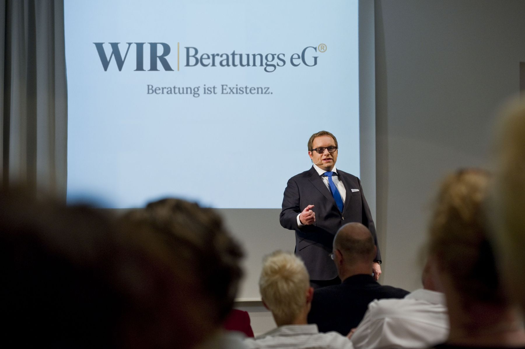 Immo Heinrich hält einen Vortrag vor Publikum, auf der Leinwand das Logo der WIR Beratungs eG